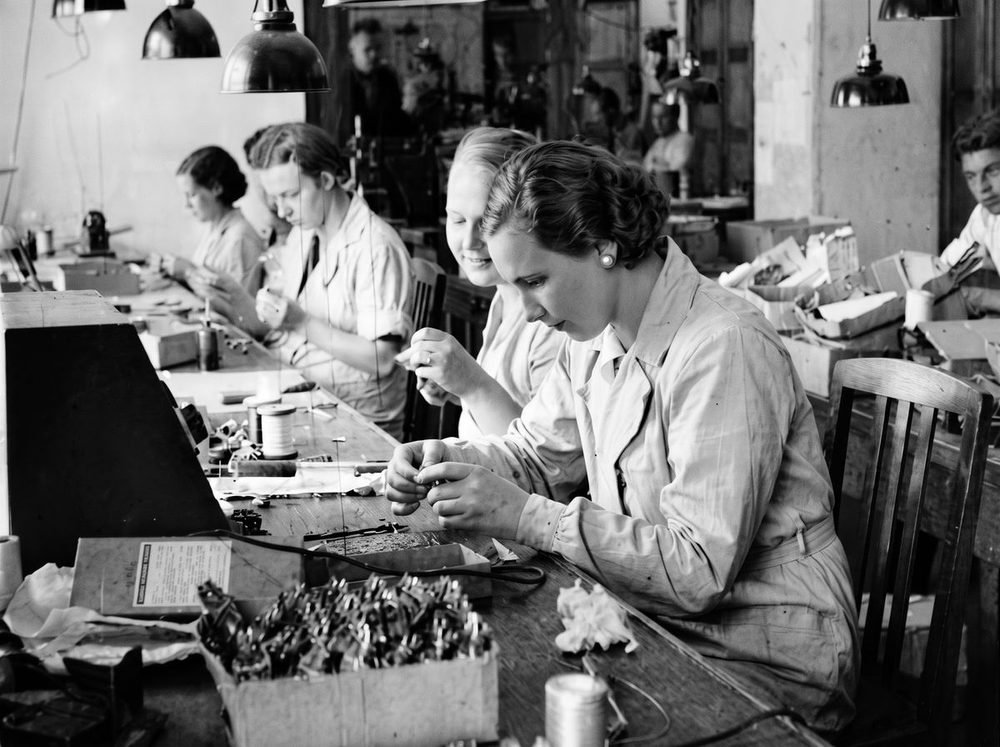 Archive photograph: workers hand-assembling components at a long workbench under pendant lamps